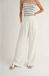 Model is wearing Sadie and Sage Heritage Wide Leg Pants, white linen pants with belt loops and pockets, relatively loose. Model is also wearing a striped tank and sandals.